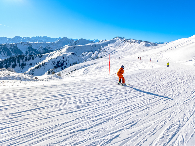 Breite verschneite Skipiste in Kitzbühel mit einem Skifahrer im orangefarbenen Outfit und Bergpanorama unter blauem Himmel.