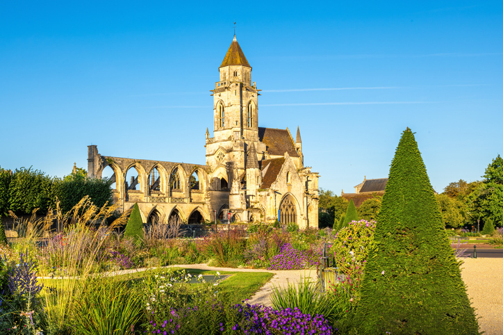 Historische Kirche in Caen mit Natursteinfassade, hohem Turm und gepflegtem Park mit Blumenbeeten und geschnittenen Sträuchern.