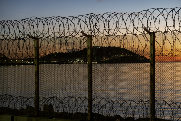 Hoher Grenzzaun mit Stacheldraht am Wasser bei Sonnenuntergang in Ceuta