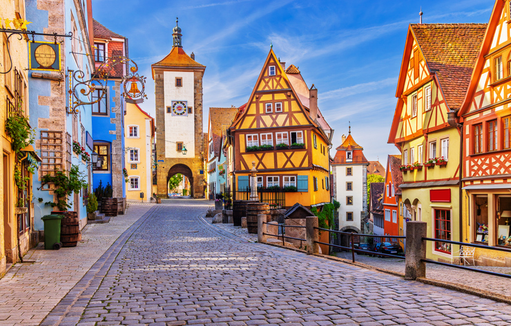 Gepflasterte Straße mit farbenfrohen Fachwerkhäusern und einem Stadttor in Rothenburg ob der Tauber unter blauem Himmel.