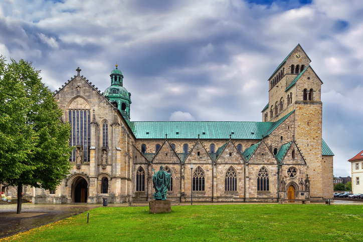 Hildesheimer Mariendom mit historischer Natursteinfassade, grünem Kupferdach, Kirchturm und Rasenfläche unter wolkigem Himmel.