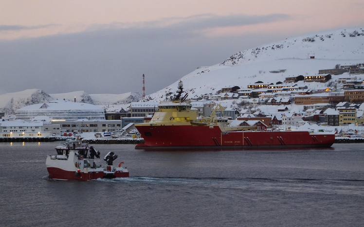 Hafen von Hammerfest im Winter mit großem rot-gelbem Schiff, kleinerem Boot, verschneiten Häusern und schneebedeckten Bergen.