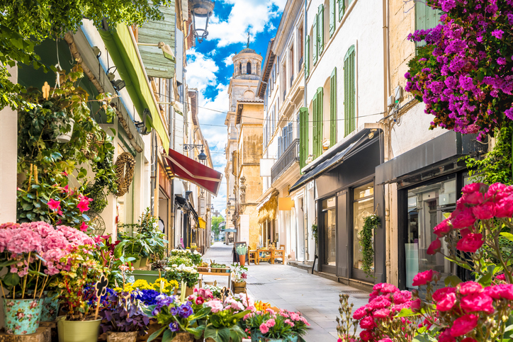 Enge Altstadtgasse in Arles mit Blumen, kleinen Geschäften und historischem Turm im Hintergrund