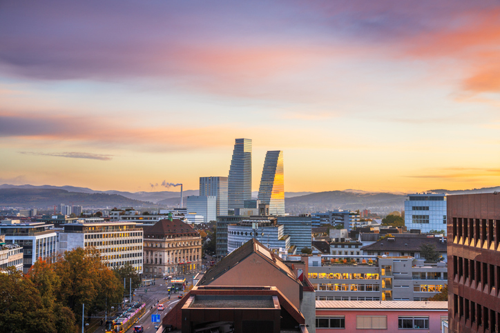 Blick über Basel bei Sonnenuntergang mit moderner Stadtsilhouette, den markanten Roche-Türmen und Bergen am Horizont.