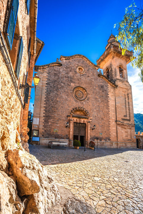 Historische Kirche Sant Bartomeu in Valldemossa mit Natursteinfassade, Rosettenfenstern und hohem Glockenturm unter blauem Himmel.
