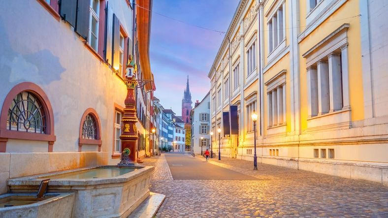 Ruhige Altstadtgasse in Basel mit Kopfsteinpflaster, historischen Häuserfassaden, beleuchteten Laternen, Brunnen und Kirchturm im Hintergrund.