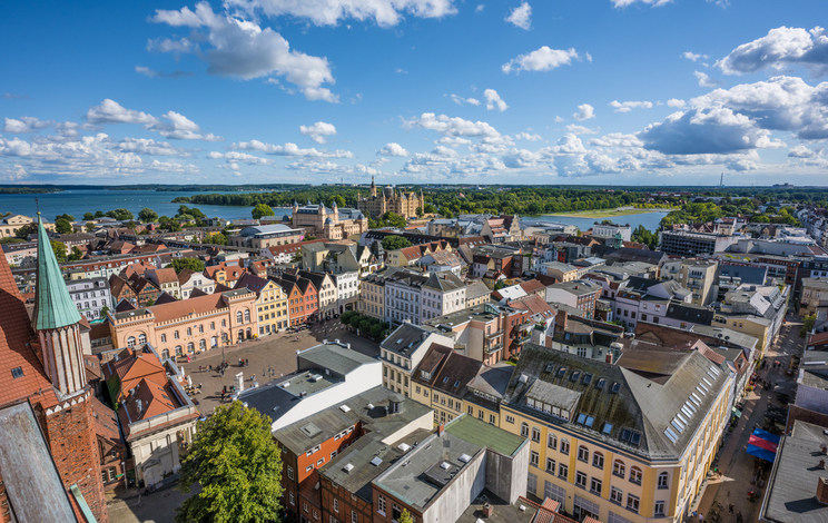 Eine Luftaufnahme zeigt die Dächer der Altstadt von Schwerin mit Kirchturm, Seen und Schloss im Hintergrund.