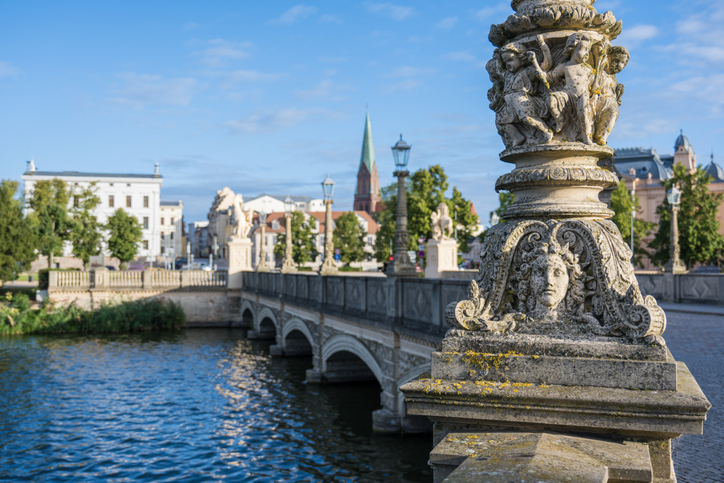 Verzierte steinerne Brückenelemente stehen im Vordergrund, dahinter spannt sich eine Brücke über das Wasser in Schwerin.