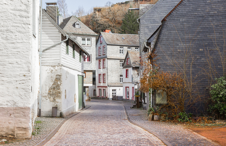 Schmale gepflasterte Gasse in Monschau mit alten Häusern, Schieferfassaden, kleinen Fenstern und steilen Hängen im Hintergrund.