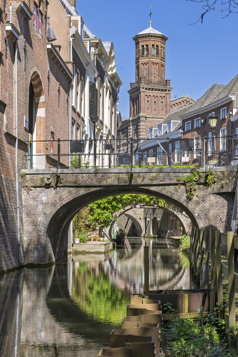 Schmale Gracht in Utrecht mit Backsteinbrücke, historischen Häusern und markantem Kirchturm unter blauem Himmel.