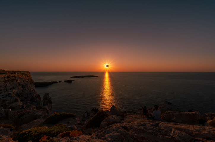 Sonnenfinsternis auf Mallorca über dem Mittelmeer bei tief stehender Sonne mit goldenem Licht auf dem Wasser und felsiger Küste im Vordergrund.