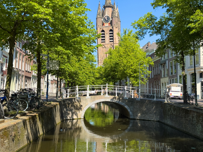 Eine kleine weiße Brücke überspannt eine Gracht in Delft, im Hintergrund ragt ein Kirchturm zwischen Bäumen auf.