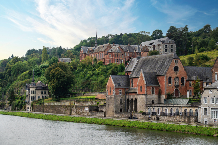 Historische Backstein- und Steinbauten am Ufer der Maas in Dinant vor grünem Hang.