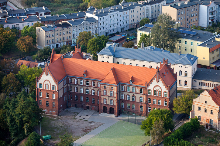 Luftaufnahme eines historischen Backsteingebäudes in Gniezno mit rotem Ziegeldach, Innenhof und Sportplatz im Vordergrund.
