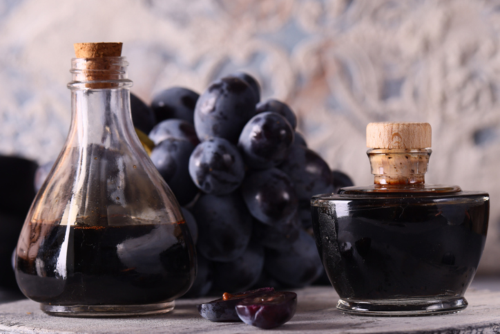 Dunkler Balsamico-Essig aus Modena in zwei Glasflaschen mit roten Weintrauben und einzelnen Traubenhälften im Hintergrund.