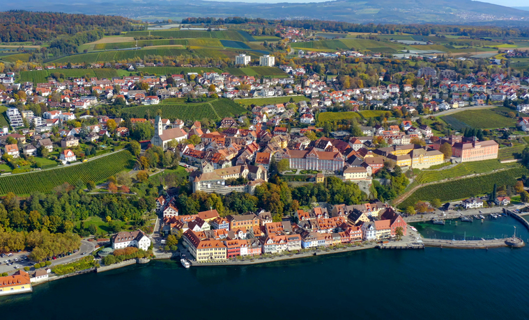Eine Luftaufnahme zeigt Meersburg mit dicht bebauter Altstadt, Weinbergen und dem Ufer des Bodensees.