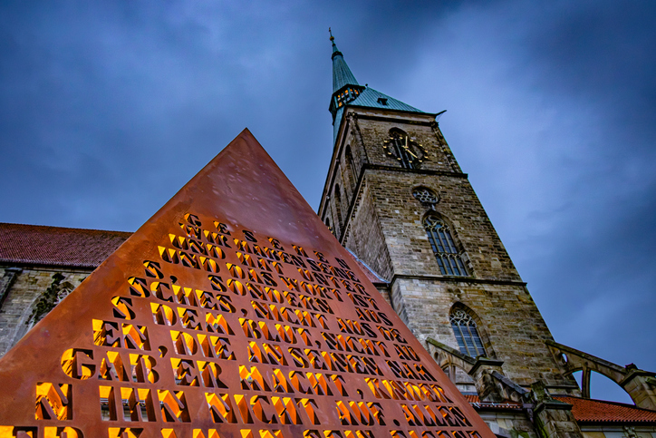 St.-Andreas-Kirche in Hildesheim mit historischem Kirchturm und beleuchteter pyramidenförmiger Skulptur des Wortes im Vordergrund bei Abendlicht.