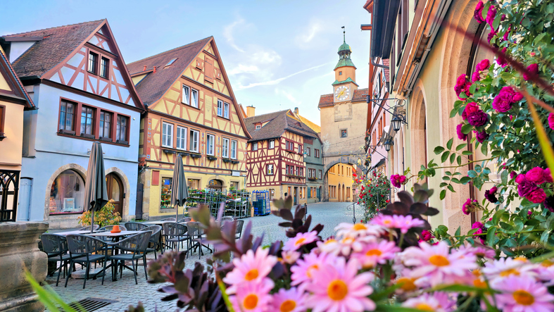 Historische Fachwerkhäuser, blühende Blumen und ein Stadttor prägen eine malerische Gasse in Rothenburg ob der Tauber.