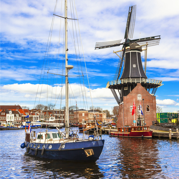 Segelboot auf dem Wasser vor der Windmühle De Adriaan in Haarlem mit historischen Gebäuden und blauem Himmel.