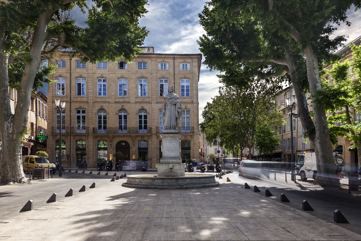 Eine steinerne Statue steht auf einem kleinen Platz in Aix-en-Provence, umgeben von Bäumen und klassischen Gebäuden.