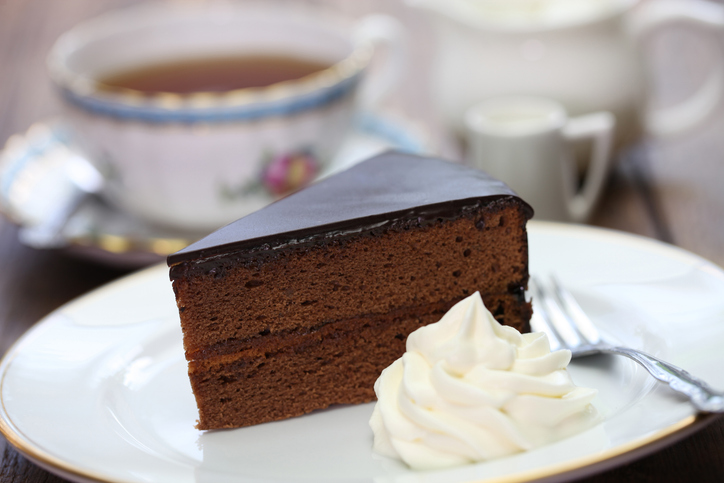 Stück Wiener Sachertorte mit glänzender Schokoladenglasur und Schlagsahne auf einem weißen Teller, dazu eine Tasse Tee im Hintergrund.