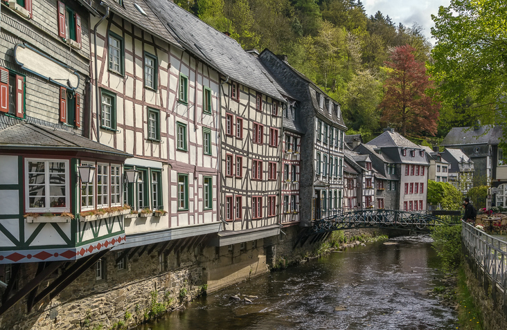 Historische Fachwerk- und Schieferhäuser in Monschau direkt an der Rur mit kleiner Brücke und grünen Bäumen am Ufer.