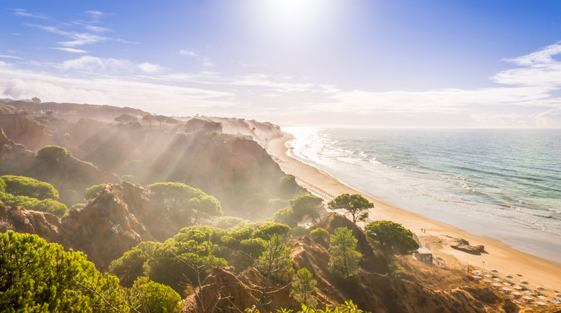 Sonniger Küstenabschnitt bei Albufeira mit langen Sandstränden, goldenen Klippen, Pinien und Lichtstrahlen über dem Meer.