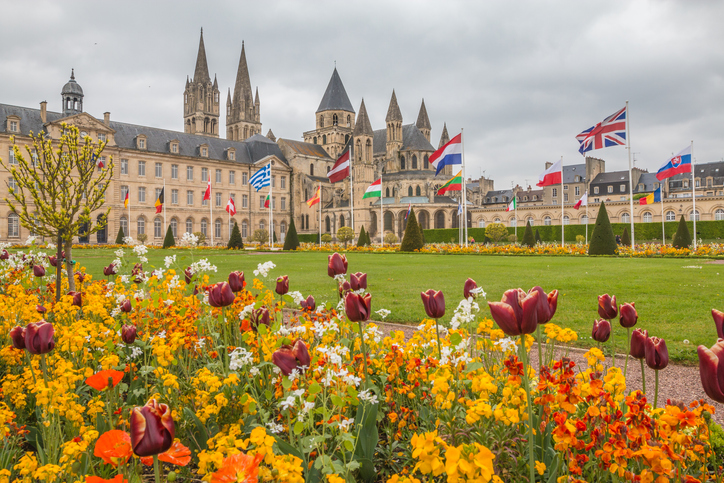 Blumenbeete und Flaggen vor einem historischen Gebäudeensemble in Caen mit gepflegter Rasenfläche und romanischen Kirchtürmen im Hintergrund.