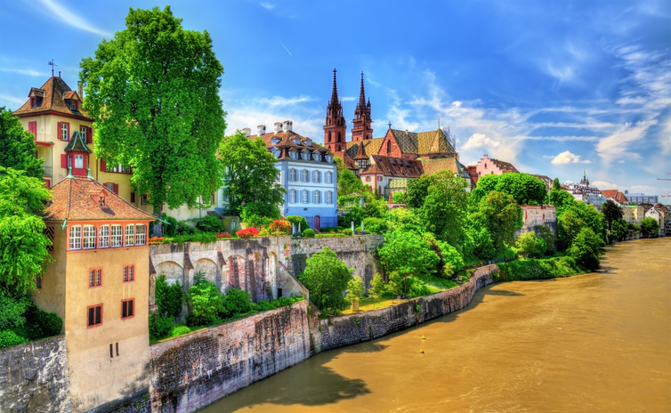 Blick auf Basel am Rhein mit grüner Uferpromenade, historischen Häusern und den beiden Türmen des Basler Münsters unter blauem Himmel.