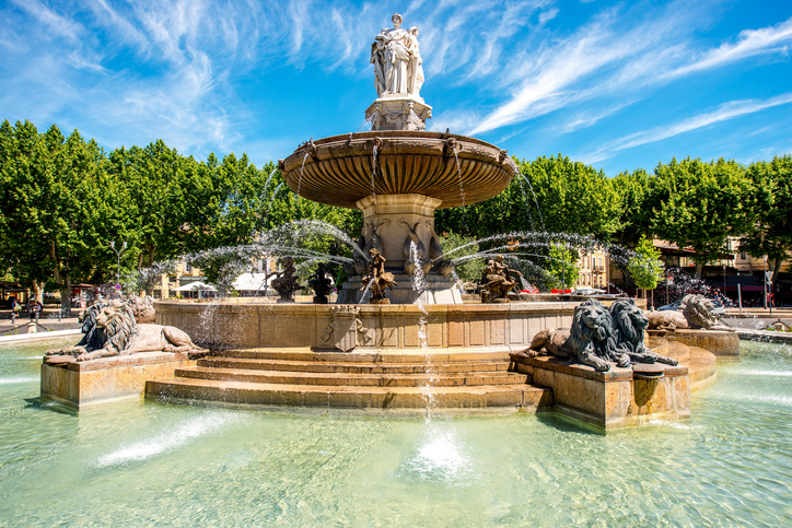Ein großer Springbrunnen mit Figuren und Wasserspielen steht auf einem sonnigen Platz in Aix-en-Provence.