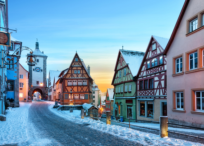 Verschneite Straße mit bunten Fachwerkhäusern und einem Torturm in Rothenburg ob der Tauber bei winterlicher Abendstimmung.