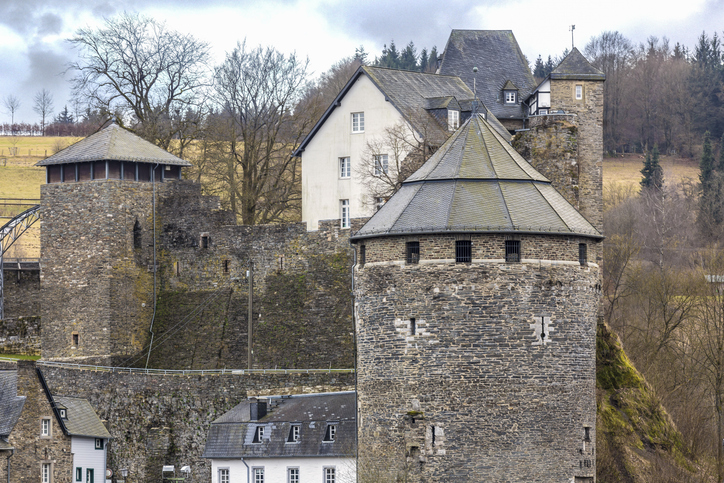 Alte Burgmauern und runde Wehrtürme der Burg Monschau oberhalb der Stadt mit Natursteinmauerwerk und ländlicher Umgebung.