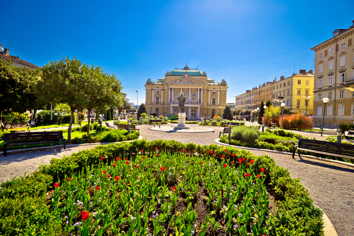 Gepflegte Parkanlage mit Blumenbeeten und Blick auf ein großes historisches Theater in Rijeka