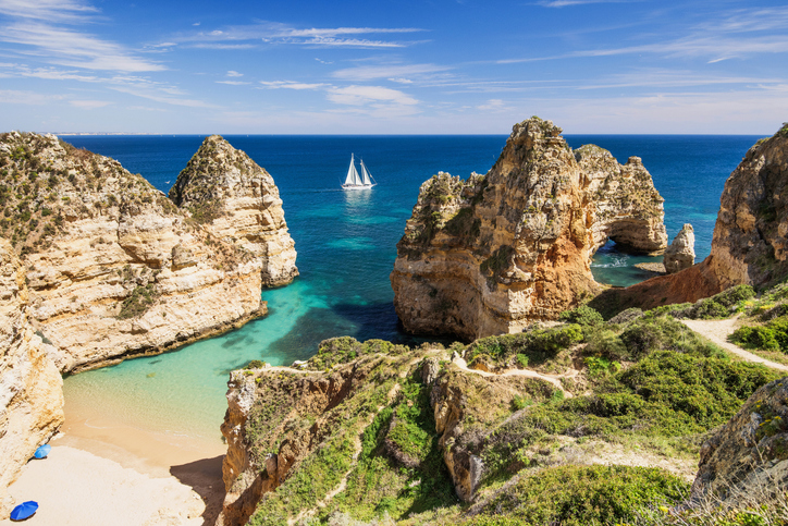 Eindrucksvolle Felsformationen bei Albufeira mit hellem Sand, türkisfarbenem Wasser, natürlichem Felsbogen und Segelboot im Hintergrund.