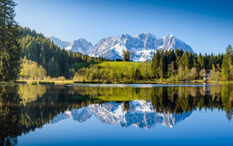 Ruhiger See bei Kitzbühel mit Spiegelung von Wäldern, Wiesen und schneebedeckten Bergen.