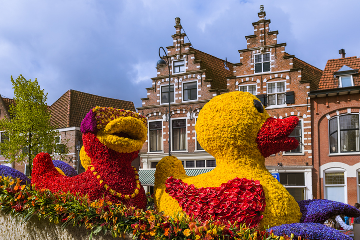 Bunte Blumenfiguren auf einem Festwagen in Haarlem vor historischen Backsteinhäusern und bewölktem Himmel.