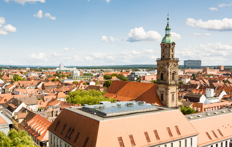 Blick über die Dächer der Erlanger Innenstadt mit historischem Kirchturm, roten Ziegeldächern und weiter Stadtlandschaft unter blauem Himmel.