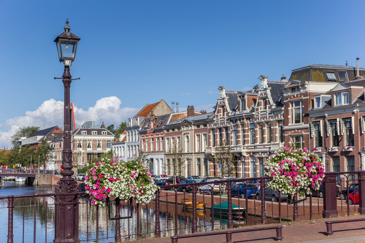 Gracht in Haarlem mit historischen Häuserfassaden, Blumenampeln an einer Brücke und ruhigem Wasser unter blauem Himmel.