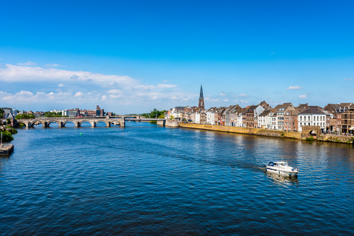 Panorama über die Maas in Maastricht mit Brücke, Uferbebauung, Kirchturm und kleinem Boot auf dem Wasser.