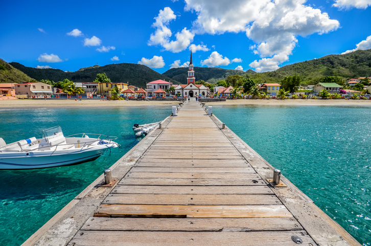 Langer Holzsteg führt über türkisfarbenes Wasser auf eine kleine Ortschaft mit Kirche und grünen Bergen auf Martinique zu.