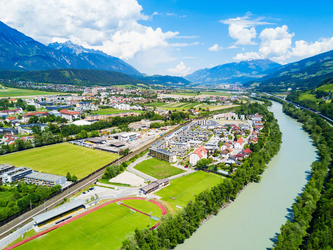 Blick über Hall in Tirol mit Fluss, Sportanlagen, Wohngebieten und Bergen unter blauem Himmel.