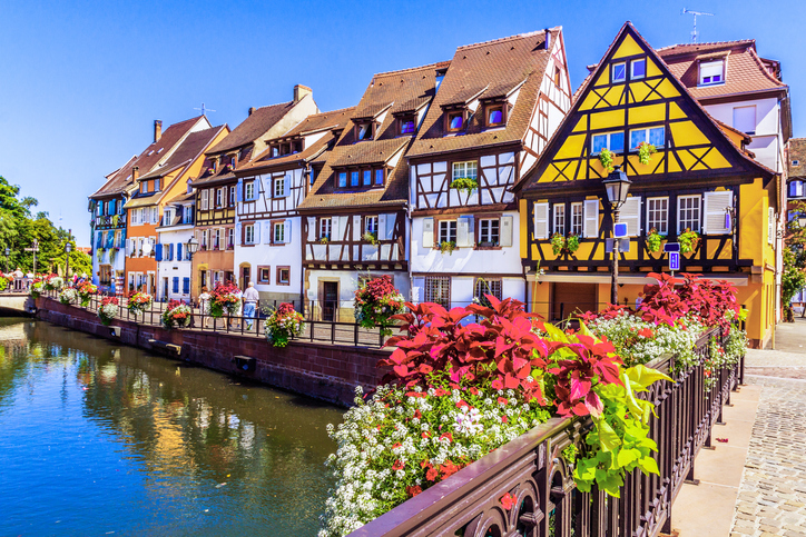 Ein Kanal mit kleiner Brücke verläuft durch Colmar, umgeben von Fachwerkhäusern und einer historischen Gebäudefassade.