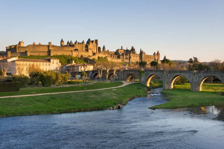 Ein Fluss und eine historische Steinbrücke führen auf die hoch über der Landschaft gelegene Festungsstadt Carcassonne zu.