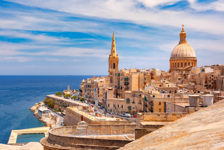 Sonniger Ausblick auf Valletta auf Malta mit dicht bebauten Natursteinhäusern, großer Kuppel, Kirchturm und tiefblauem Mittelmeer.
