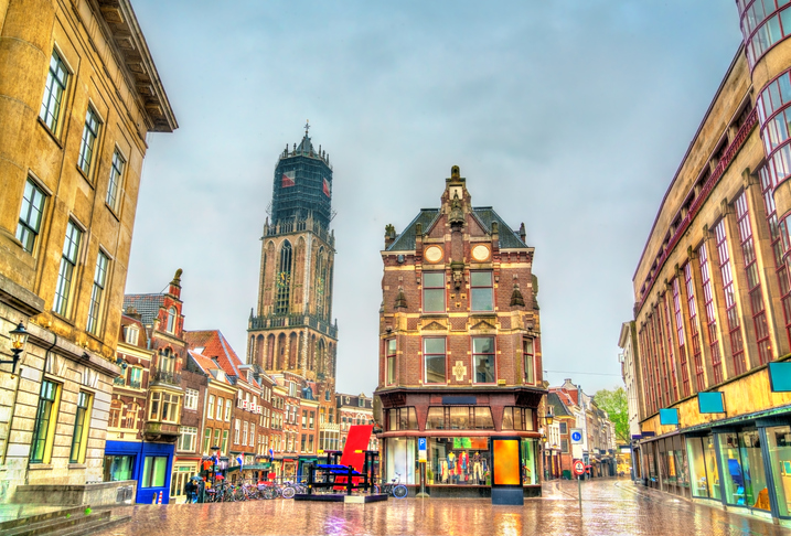 Historischer Platz in Utrecht mit Backstein- und Giebelhäusern sowie dem Domturm im Hintergrund unter bewölktem Himmel.