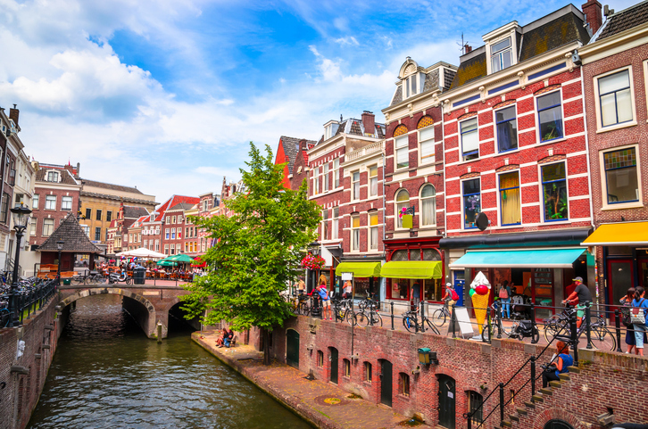 Belebte Oudegracht in Utrecht mit historischen Backsteinhäusern, Geschäften, Fahrrädern, Brücke und tiefer liegendem Kanal.