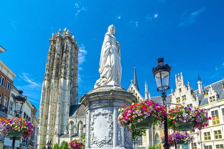 Historische Statue auf einem hohen Sockel in Mechelen mit St.-Rumbold-Kathedrale, Blumenampeln und blauem Himmel im Hintergrund.